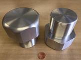 An up close comparison of two machined aluminum threaded plugs standing next to a penny